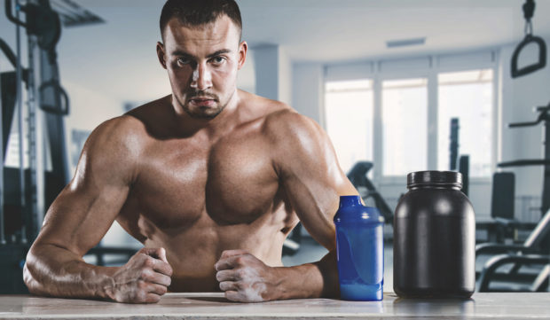 Muscular man sitting sweating in gym with drinking cup and supplement.